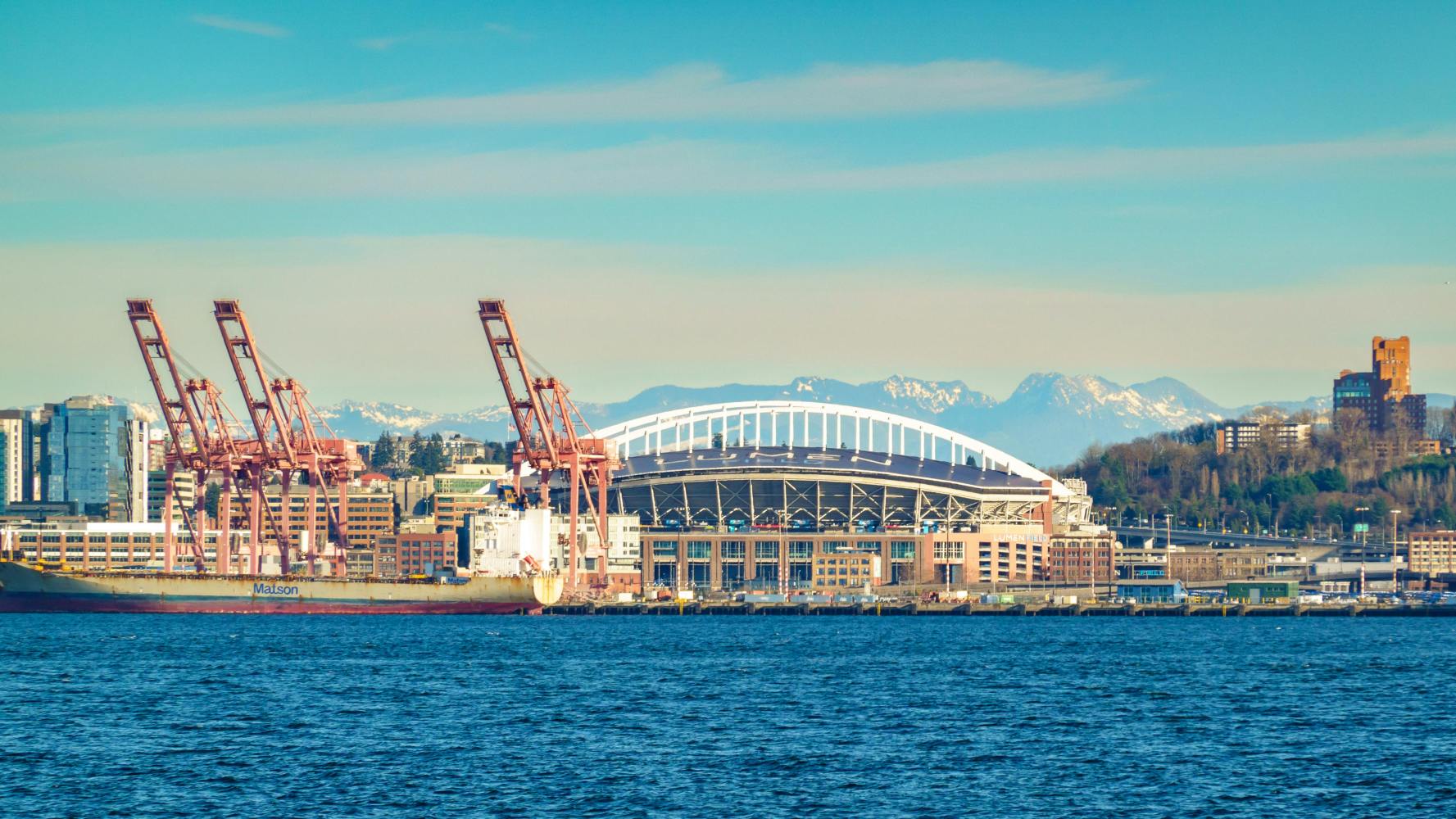 Stadium near waterfront with cranes and mountains in background.